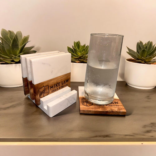 Wooden business card holder with 'MIKE LAW Injury Attorney' branding, marble coasters, and a glass of water on a wooden surface with plants in the background.